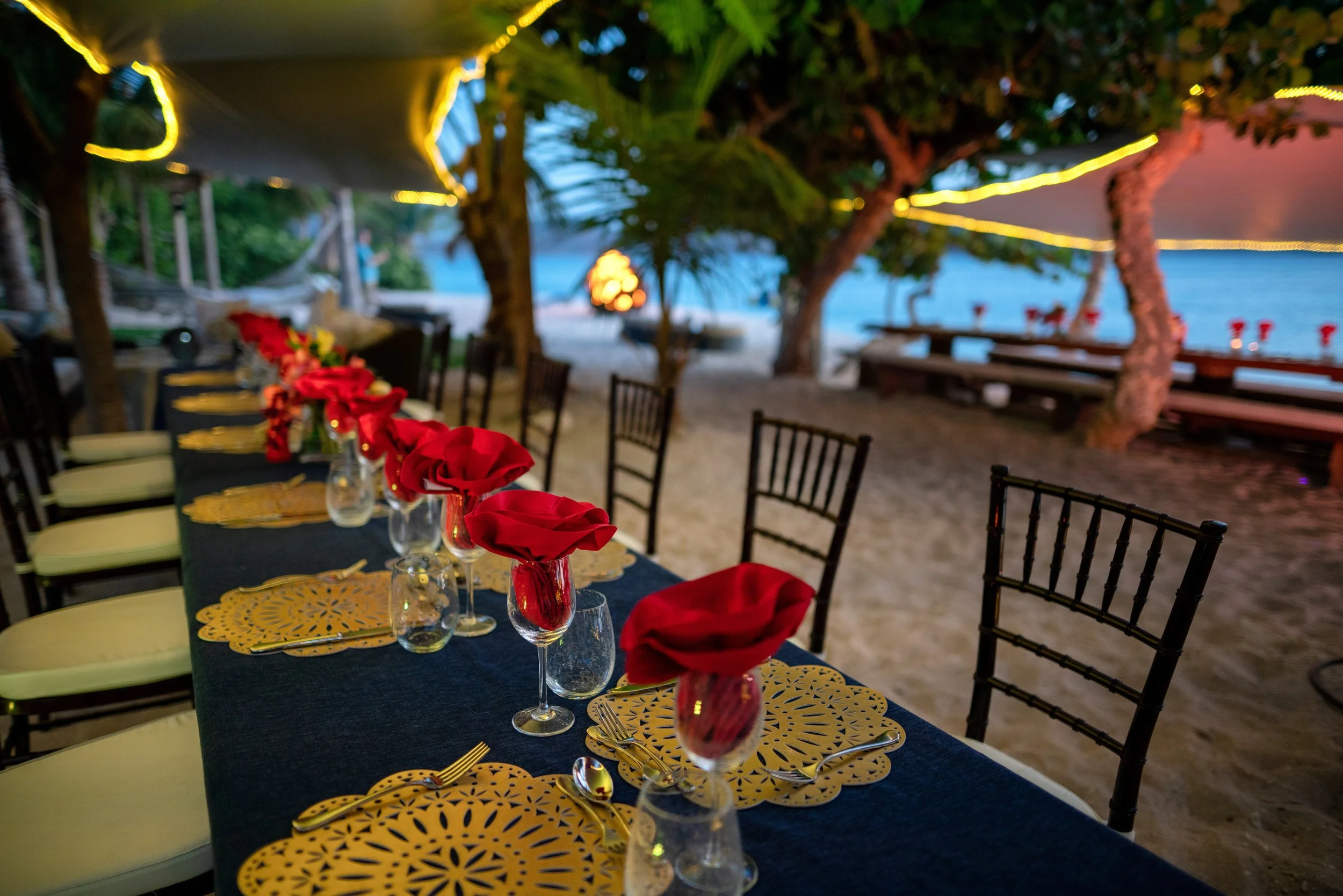 A long dinner table laid on the sand at dusk, with red napkins and lights strung through the trees